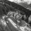 A winding mountain road with sharp curves cuts through a forested slope; snow patches line the road and distant snow-covered peaks are visible in the background. Nature and landscape poster print available in A1 to A4 sizes.