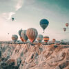 Several hot air balloons float above rocky cliffs and valleys under a partly cloudy sky, as people at the Balloon Festival observe from a viewpoint below. Nature and landscape poster print available in A1 to A4 sizes.
