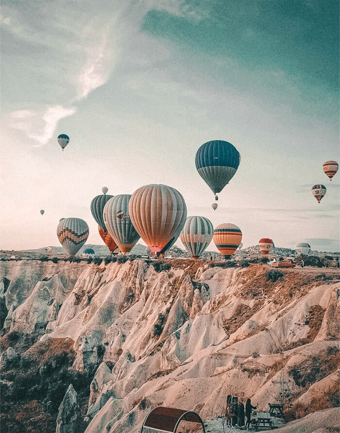 product-image-11-14 Several hot air balloons float above rocky cliffs and valleys under a partly cloudy sky, as people at the Balloon Festival observe from a viewpoint below. Nature and landscape poster print available in A1 to A4 sizes.