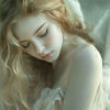 A young woman with long, wavy blonde hair and white feathered wings looks downward with a serene expression, dressed in a soft, flowing garment. Fantasy art poster available in A1 to A4 sizes.