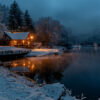 A snow-covered cabin with warm lights is reflected in a calm lake at dusk, surrounded by trees and distant lights under a cloudy, wintry sky. Wall art print available in A1 to A4 sizes.