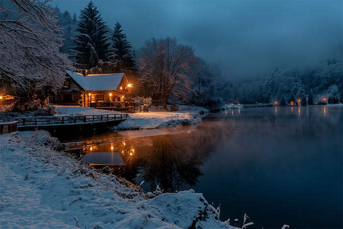 A snow-covered cabin with warm lights is reflected in a calm lake at dusk, surrounded by trees and distant lights under a cloudy, wintry sky. Wall art print available in A1 to A4 sizes.