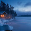 A snow-covered cabin with warmly lit windows sits beside a frozen lake and pine trees at dusk under a cloudy sky. Wall art print available in A1 to A4 sizes.