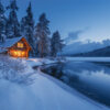 A log cabin with lit windows sits beside a snow-covered riverbank and pine trees under a twilight sky. Wall art print available in A1 to A4 sizes.