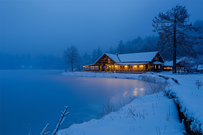 product-image-3-2 A snow-covered cabin with warm lights sits beside a frozen lake, surrounded by trees, under a blue evening sky. Wall art print available in A1 to A4 sizes.