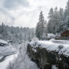 A wooden cabin sits on a snow-covered cliff overlooking a winding river, surrounded by dense, snow-laden evergreen trees. Wall art print available in A1 to A4 sizes.