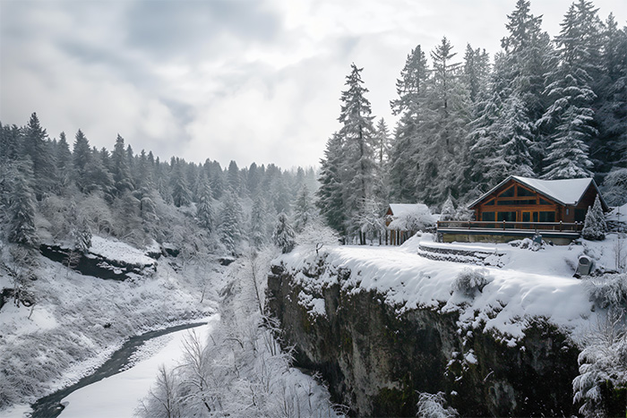 A wooden cabin sits on a snow-covered cliff overlooking a winding river, surrounded by dense, snow-laden evergreen trees. Wall art print available in A1 to A4 sizes.