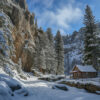 A small wooden cabin sits among snow-covered trees and rocky cliffs under a clear blue sky in a mountainous winter landscape. Wall art print available in A1 to A4 sizes.