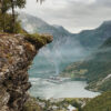 A cruise ship sails through a fjord surrounded by steep, green mountains, offering a breathtaking mountain view from a rocky cliff above a small village. Nature and landscape poster print available in A1 to A4 sizes.
