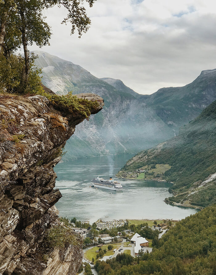 product-image A cruise ship sails through a fjord surrounded by steep, green mountains, offering a breathtaking mountain view from a rocky cliff above a small village. Nature and landscape poster print available in A1 to A4 sizes.