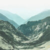 A misty mountain valley with green hills and winding paths, surrounded by layered, forested peaks under a pale sky. Nature and landscape poster print available in A1 to A4 sizes.