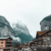 A scene from a Mountain Village unfolds: wooden buildings line wet roads, while misty mountains loom in the background beneath an overcast sky. Nature and landscape poster print available in A1 to A4 sizes.