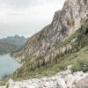 Evergreen trees descend a rocky mountain slope to the tranquil waters of a Mountain Lake, beneath a cloudy sky with distant peaks in the background. Nature and landscape poster print available in A1 to A4 sizes.