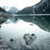 Crystal-clear Mountain Lake with rocks visible underwater in the foreground, surrounded by mountains whose snow-capped peaks are reflected in the still water. Nature and landscape poster print available in A1 to A4 sizes.