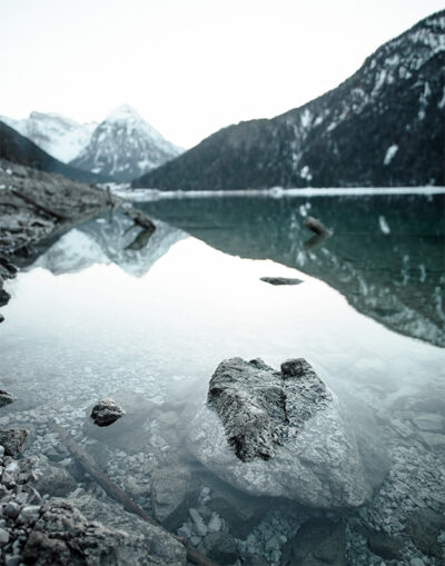 Crystal-clear Mountain Lake with rocks visible underwater in the foreground, surrounded by mountains whose snow-capped peaks are reflected in the still water. Nature and landscape poster print available in A1 to A4 sizes.