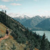 Two hikers walk along a Mountain Path, a narrow hiking trail bordered by pine trees, with a lake below and snow-capped mountains rising in the distance under a partly cloudy sky. Nature and landscape poster print available in A1 to A4 sizes.
