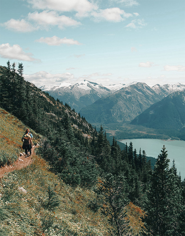 product-image Two hikers walk along a Mountain Path, a narrow hiking trail bordered by pine trees, with a lake below and snow-capped mountains rising in the distance under a partly cloudy sky. Nature and landscape poster print available in A1 to A4 sizes.