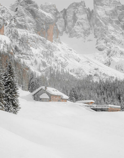 A snow-covered mountain lodge stands among pine trees with large, rocky mountains in the background under a cloudy sky. Nature and landscape poster print available in A1 to A4 sizes.