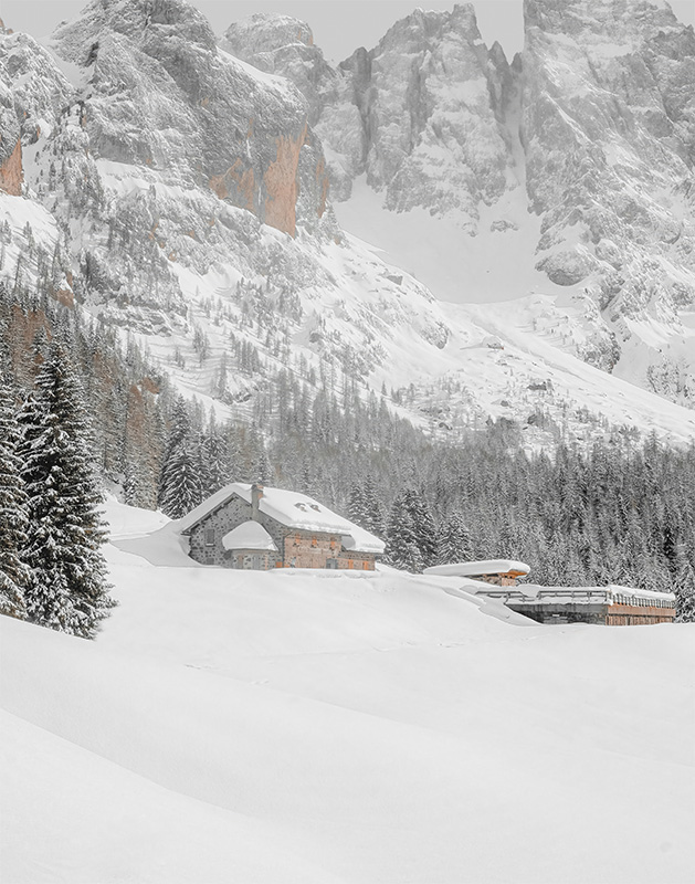 product-image A snow-covered mountain lodge stands among pine trees with large, rocky mountains in the background under a cloudy sky. Nature and landscape poster print available in A1 to A4 sizes.