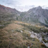 Rocky mountain landscape with grassy foreground, scattered stones, and rugged peaks rising beyond a scenic mountain pass under a partly cloudy sky. Nature and landscape poster print available in A1 to A4 sizes.