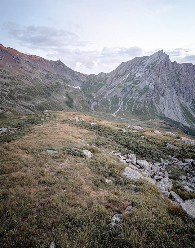 product-image Rocky mountain landscape with grassy foreground, scattered stones, and rugged peaks rising beyond a scenic mountain pass under a partly cloudy sky. Nature and landscape poster print available in A1 to A4 sizes.