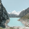 A turquoise Mountain Lake stretches between rocky mountains, with snow-capped peaks in the distance beneath a clear sky. A small boat and kayak rest on the shore. Nature and landscape poster print available in A1 to A4 sizes.