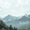 Snow-capped peaks of Mountain Range rise above a forested valley under a cloudy sky, with mist partially covering the lower slopes. Nature and landscape poster print available in A1 to A4 sizes.