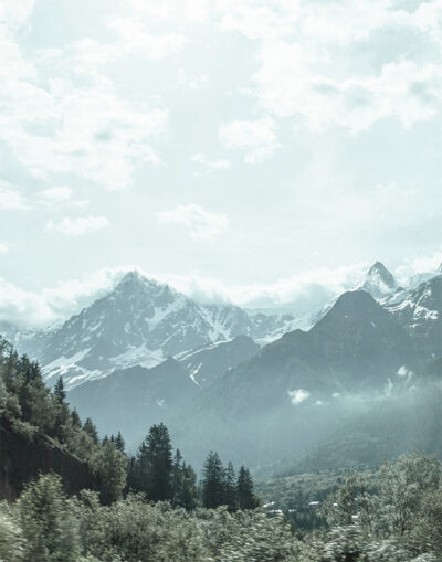 Snow-capped peaks of Mountain Range rise above a forested valley under a cloudy sky, with mist partially covering the lower slopes. Nature and landscape poster print available in A1 to A4 sizes.