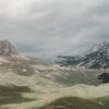 A wide, grassy valley with rolling foothills and rocky mountains beneath a cloudy sky. Nature and landscape poster print available in A1 to A4 sizes.