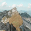 A rugged mountain peak with grassy slopes and rocky ridges rises under a partly cloudy sky, reminiscent of Mountain Ridge scenery, and is surrounded by distant mountain ranges. Nature and landscape poster print available in A1 to A4 sizes.