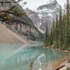 A clear turquoise mountain lake reflects pine trees and a snow-capped mountain, with rocky slopes and cloudy skies in the background. Nature and landscape poster print available in A1 to A4 sizes.