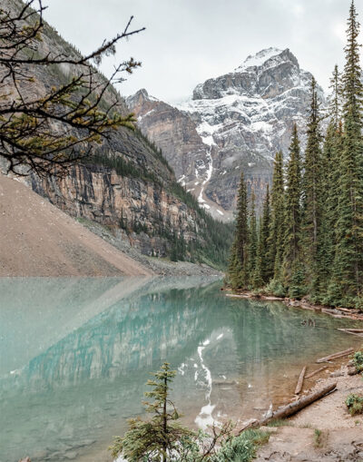 A clear turquoise mountain lake reflects pine trees and a snow-capped mountain, with rocky slopes and cloudy skies in the background. Nature and landscape poster print available in A1 to A4 sizes.
