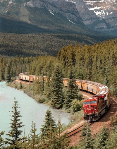 A red Mountain Train pulls a long freight train around a curve, passing through dense forest alongside a river with mountains in the background. Nature and landscape poster print available in A1 to A4 sizes.