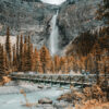 A tall waterfall cascades down a rocky cliff in the background; a metal footbridge crosses a river in the foreground, surrounded by trees with vibrant autumn foliage. Nature and landscape poster print available in A1 to A4 sizes.