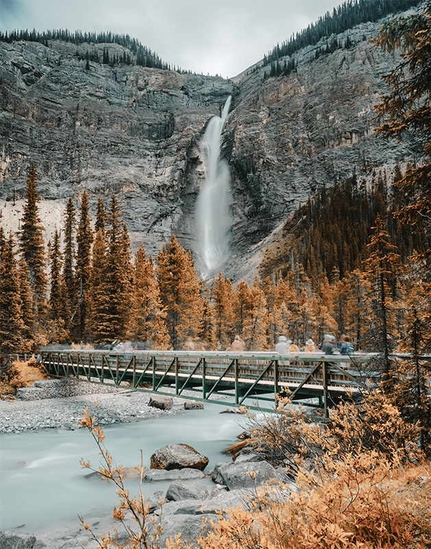 product-image A tall waterfall cascades down a rocky cliff in the background; a metal footbridge crosses a river in the foreground, surrounded by trees with vibrant autumn foliage. Nature and landscape poster print available in A1 to A4 sizes.