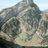 Steep mountain with sharp peak rises above a grassy valley, where a mountain village with scattered houses and trees nestles at its base under a clear blue sky. Nature and landscape poster print available in A1 to A4 sizes.