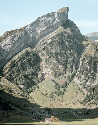 Steep mountain with sharp peak rises above a grassy valley, where a mountain village with scattered houses and trees nestles at its base under a clear blue sky. Nature and landscape poster print available in A1 to A4 sizes.