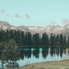 A calm mountain lake reflects evergreen trees and distant snow-capped mountains under a clear blue sky. Nature and landscape poster print available in A1 to A4 sizes.