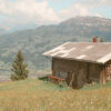 A mountain cabin sits on a grassy hillside with wildflowers, overlooking distant peaks under a partly cloudy sky—the perfect mountain getaway. Nature and landscape poster print available in A1 to A4 sizes.