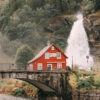 A red house sits beside a stone bridge and river, with the impressive Steinsdalsfossen Waterfall and dense trees in the background, capturing the scenic beauty of Norway. Nature and landscape poster print available in A1 to A4 sizes.