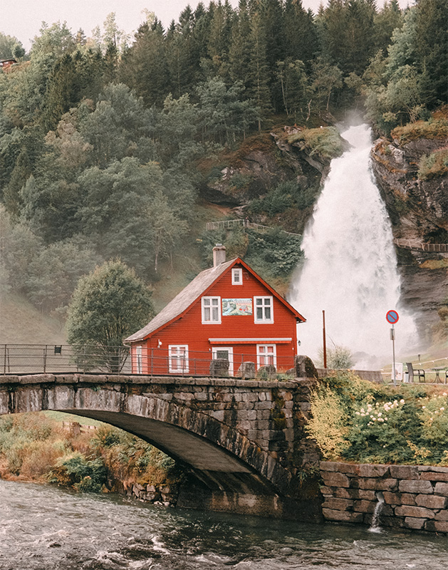 product-image A red house sits beside a stone bridge and river, with the impressive Steinsdalsfossen Waterfall and dense trees in the background, capturing the scenic beauty of Norway. Nature and landscape poster print available in A1 to A4 sizes.