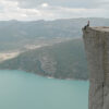 A person sits at the edge of a high cliff overlooking a turquoise lake and distant mountains under a cloudy sky. Nature and landscape poster print available in A1 to A4 sizes.