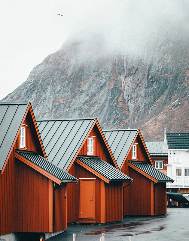 Three red wooden cabins with steep roofs stand in front of a misty mountain in Reine, Norway, with a bird flying in the cloudy sky above. Nature and landscape poster print available in A1 to A4 sizes.
