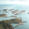 A coastal landscape in Norway, featuring the Lofoten Islands with small islets connected by winding roads and bridges, surrounded by a calm blue sea under a partly cloudy sky. Nature and landscape poster print available in A1 to A4 sizes.