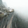 A Man On A Ledge holding an umbrella stands near the edge of a steep, rocky cliff in a foggy, mountainous landscape. Nature and landscape poster print available in A1 to A4 sizes.