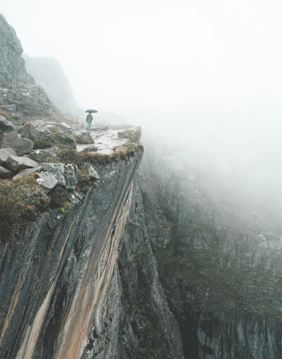 A Man On A Ledge holding an umbrella stands near the edge of a steep, rocky cliff in a foggy, mountainous landscape. Nature and landscape poster print available in A1 to A4 sizes.