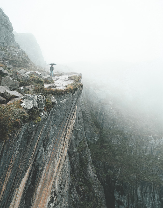 A Man On A Ledge holding an umbrella stands near the edge of a steep, rocky cliff in a foggy, mountainous landscape. Nature and landscape poster print available in A1 to A4 sizes.