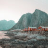 Red cabins on a rocky shoreline near Leknes, Norway, with large mountains rising in the background beneath a pale sky. Nature and landscape poster print available in A1 to A4 sizes.