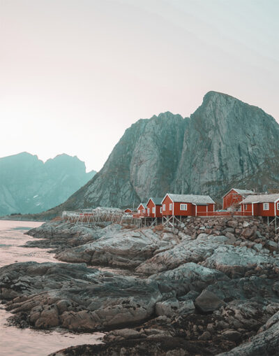 Red cabins on a rocky shoreline near Leknes, Norway, with large mountains rising in the background beneath a pale sky. Nature and landscape poster print available in A1 to A4 sizes.