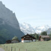 A brown wooden house stands in a grassy field with a fence in front, surrounded by Switzerland’s mountains and snowy peaks under a clear sky. Nature and landscape poster print available in A1 to A4 sizes.